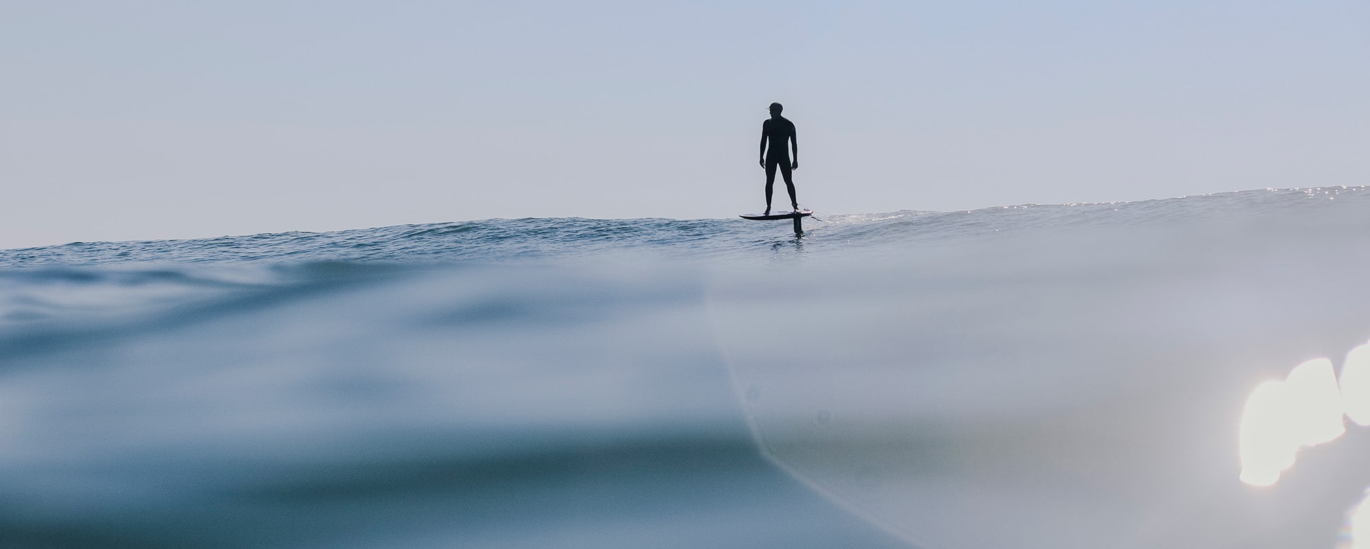 A solitary figure stands atop a hydrofoil board, equipped with a Cedrus Evolution Wind mast, gliding across the calm ocean surface. The rider's relaxed silhouette contrasts with the clear sky, evoking the tranquility and balance of foiling. The mast, slicing through the water, promises high performance and stability even in such serene conditions.