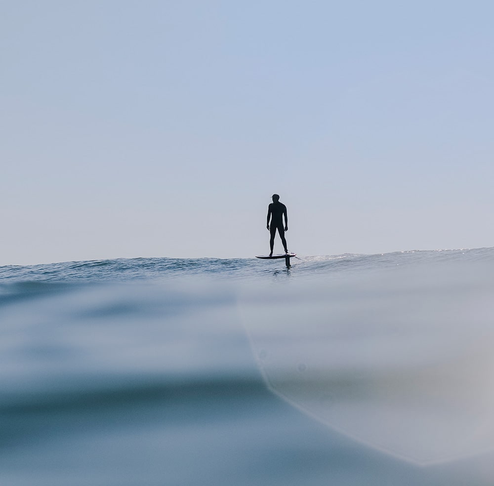A solitary figure stands atop a hydrofoil board, equipped with a Cedrus Evolution Wind mast, gliding across the calm ocean surface. The rider's relaxed silhouette contrasts with the clear sky, evoking the tranquility and balance of foiling. The mast, slicing through the water, promises high performance and stability even in such serene conditions.