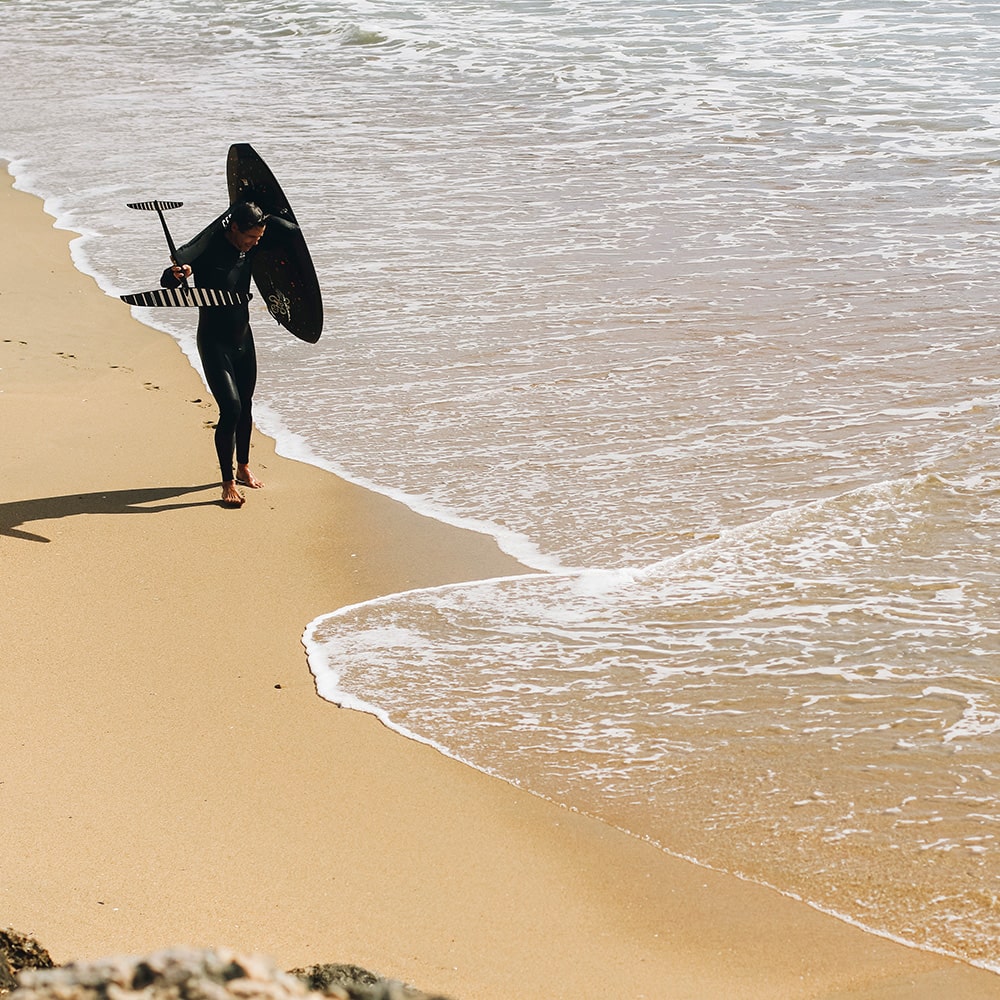 Walking along the shoreline, a surfer carries a Cedrus mast and hydrofoil board un, ready to answer the ocean's call. The sandy beach and foamy waves set the stage for a day of gliding over the water, encapsulating the anticipation and preparation that goes hand-in-hand with the sport. This moment, symbolic of the pre-ride ritual, serves as the perfect visual introduction to the frequently asked questions about hydrofoiling with Cedrus.