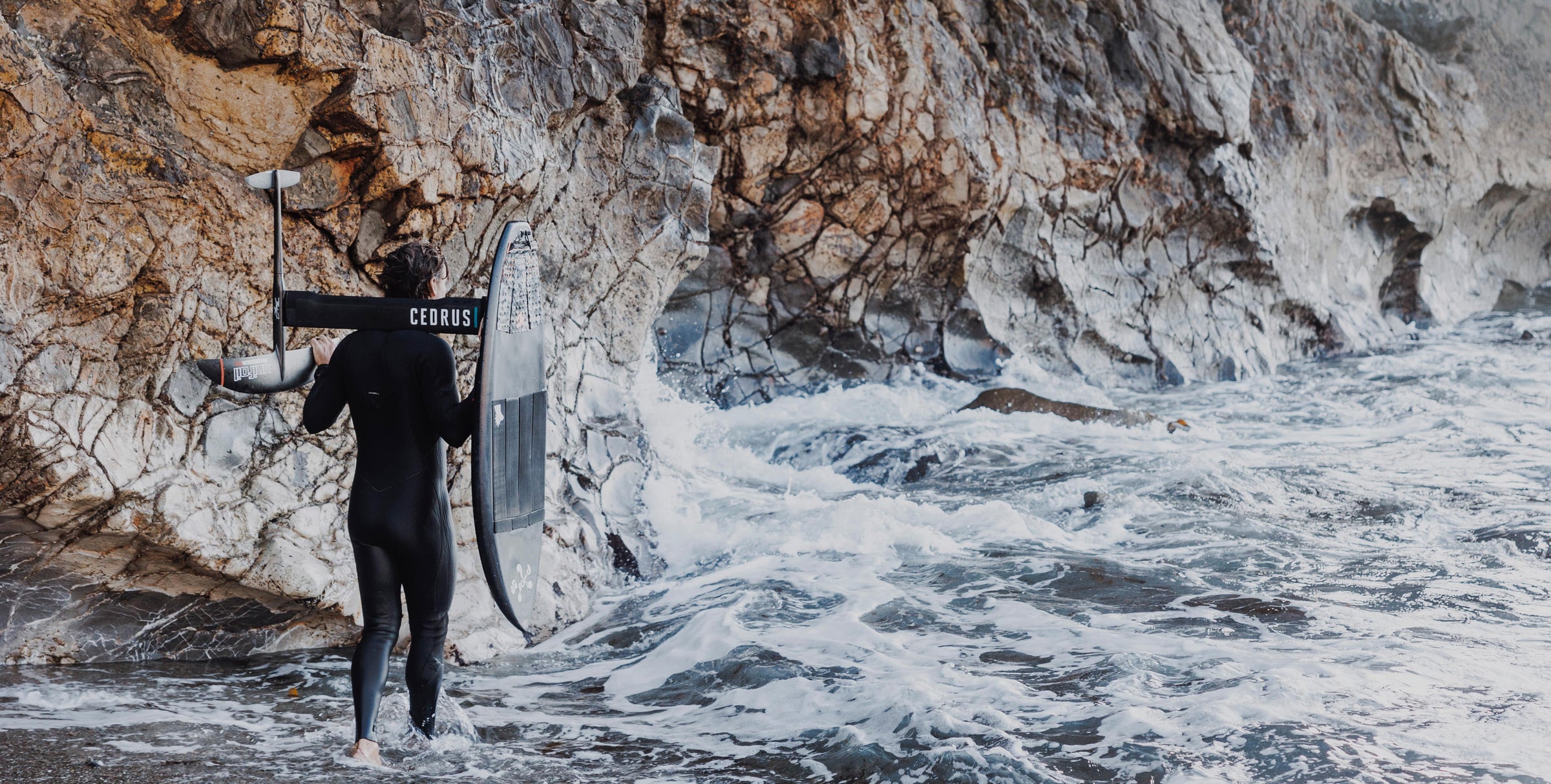 Image with surfer walking out to the ocean against a large cliff. Cedrus masts are custom made, high performance masts for any foil system.