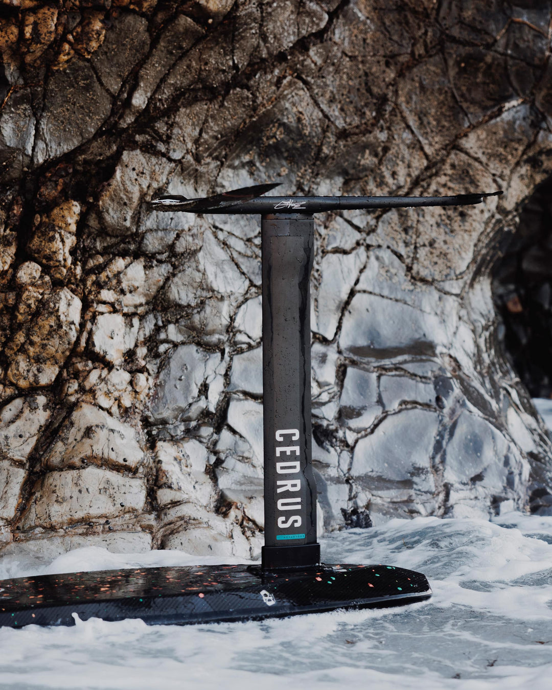 Cedrus hydrofoil mast mounted on a board float against a backdrop of smooth and seemingly polished rocks, its sustainable engineering on full display. The carbon fiber mast, with its logo, speaks to meticulous design and manufacturing that values precision and minimal waste. The hollow core construction of the mast is a testament to efficiency, reducing material use without compromising performance, reflecting Cedrus' commitment to responsible production practices in hydrofoil technology.
