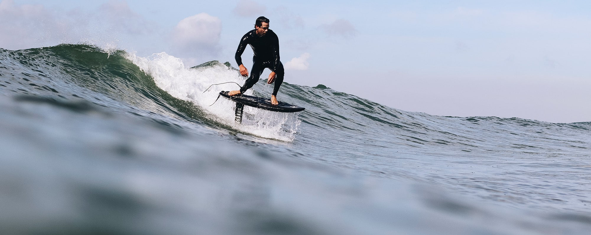 Rider draws a clean line as he drops into a wave at the moment the board is rising out of the water from the lift of the foil and water can be seen running off of the deck of the board. The rider is confidently balanced on a hydrofoil equipped with the Cedrus Evolution Surf Mast. The mast's sleek profile cuts through the water's surface, a testament to Cedrus' commitment to elevating the foiling experience through advanced design and engineering.