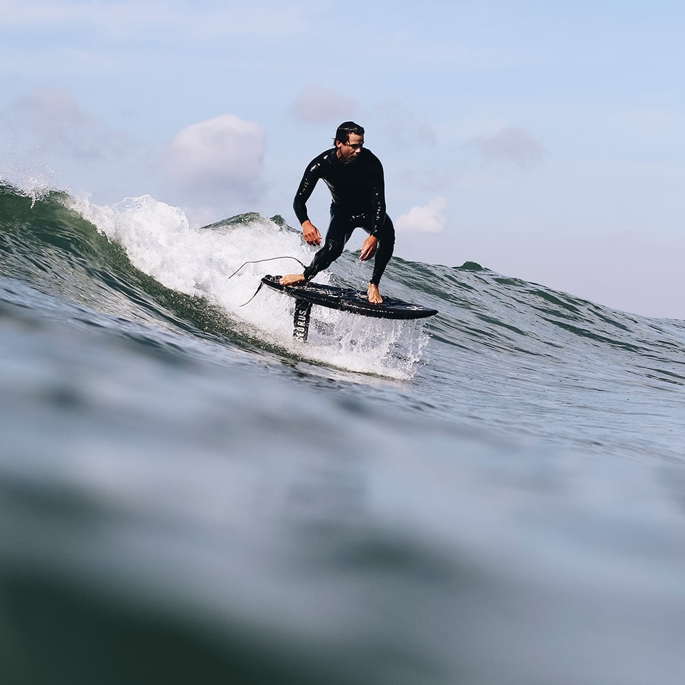 Rider draws a clean line as he drops into a wave at the moment the board is rising out of the water from the lift of the foil and water can be seen running off of the deck of the board. The rider is confidently balanced on a hydrofoil equipped with the Cedrus Evolution Surf Mast. The mast's sleek profile cuts through the water's surface, a testament to Cedrus' commitment to elevating the foiling experience through advanced design and engineering.