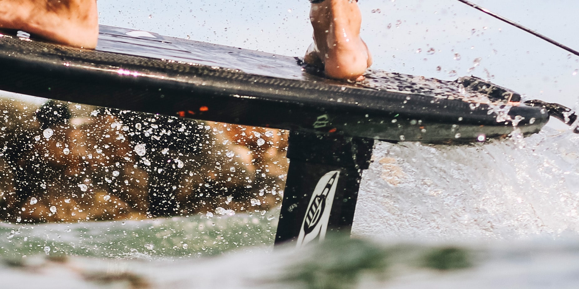 Close-up view of a hydrofoil board riding a wave, focusing on the Cedrus Evolution Mast attached beneath the board. The mast, known for its advanced engineering and unique water-piercing design, cuts through the water with precision, highlighted by the shimmering water droplets in mid-air. This hydrofoil is tailored for enthusiasts who demand both performance and innovation in their foiling experience.