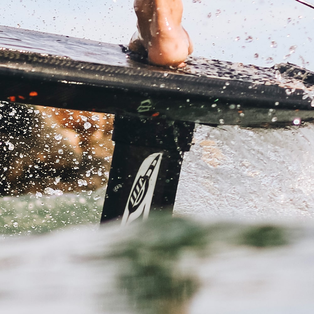 Close-up view of a hydrofoil board riding a wave, focusing on the Cedrus Evolution Mast attached beneath the board. The mast, known for its advanced engineering and unique water-piercing design, cuts through the water with precision, highlighted by the shimmering water droplets in mid-air. This hydrofoil is tailored for enthusiasts who demand both performance and innovation in their foiling experience.