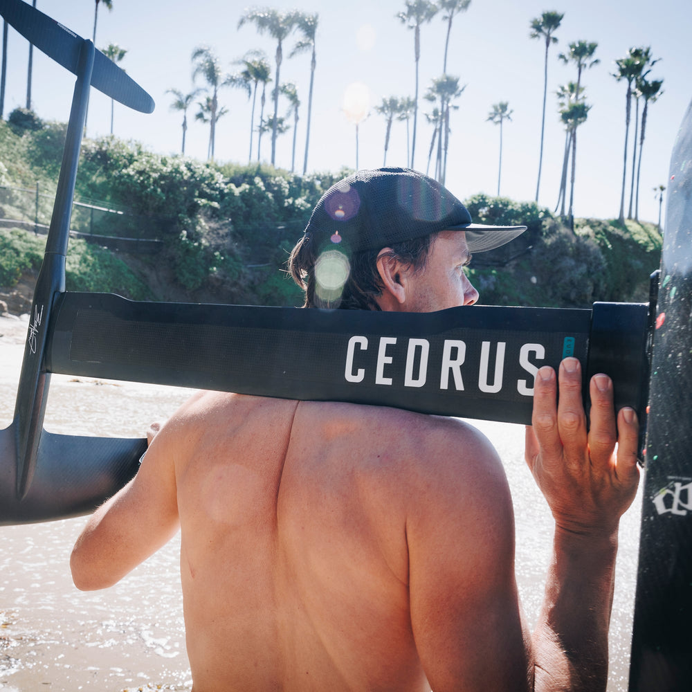 A foiler carries the Cedrus Evolution Surf Mast over their shoulder, with its signature branding visible, against a sunny beach backdrop lined with palm trees, reflecting the mast's seamless integration into the foiling lifestyle