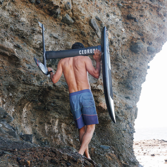 A surfer walks on rocks for a session, holding the Cedrus Evolution Surf Mast and board against a rugged cliffside, showcasing the equipment's durability and the adventurous spirit it embodies.