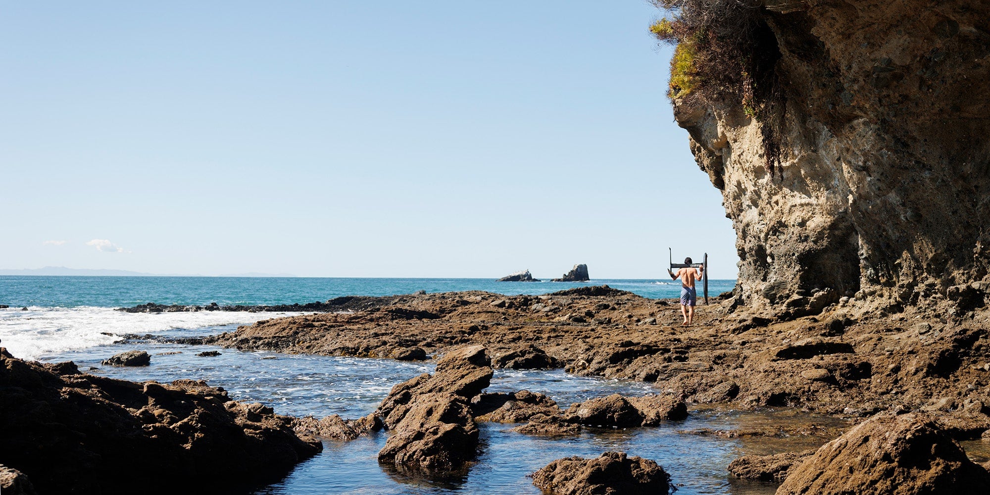 A serene coastal scene with a person standing on rocky tidal flats, holding a hydrofoil surfboard equipped with a Cedrus Evolution Surf Mast above their head. The individual is facing the ocean, seemingly ready to jump into the ocean. In the background, the calm blue sea stretches to the horizon, dotted with small islets. A prominent weathered cliff to the right towers over the area, contributing to the peaceful yet wild atmosphere of this picturesque location.