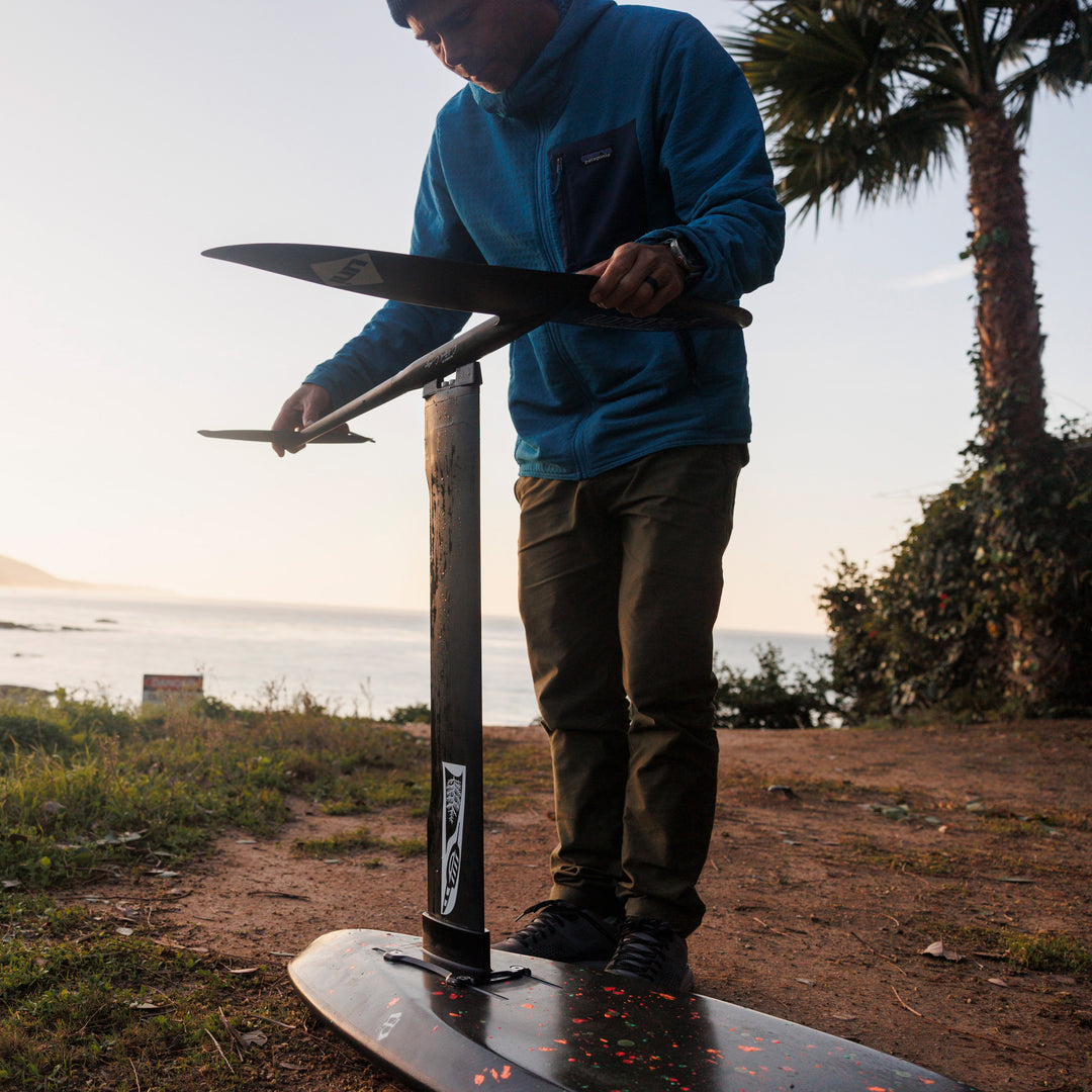 Person standing on a cliff above the ocean at sunrise installing a Unifoil foil onto a Cedrus Evolution mast with a Unifoil adapter designed an engineered by Cedrus to fit seamlessly with limitless possibilities to surf any foil brand with a Cedrus mast