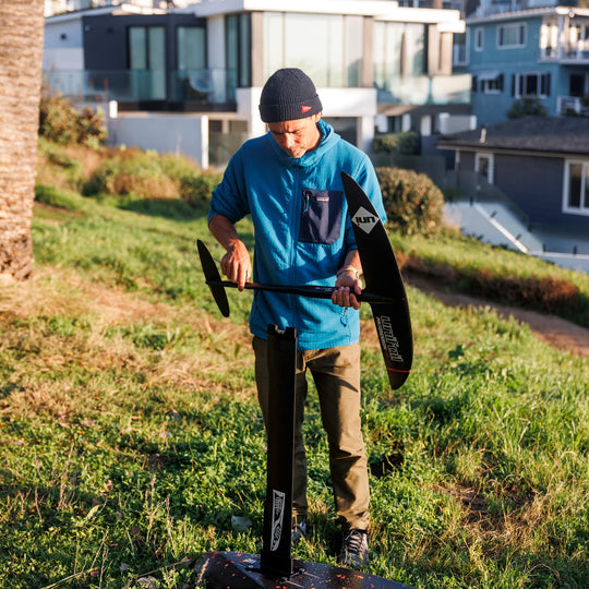 A person assembling a Unifoil setup on a Cedrus Evolution Surf mast using a Cedrus Foil Adapter for the brand of Unifoil foils, highlighting its sleek black design and robust construction with dual stainless steel posts for secure mounting. It exemplifies Cedrus' commitment to versatile, universal and seamless foil compatibility and advanced engineering