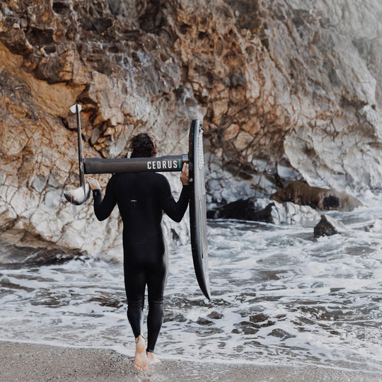 The Cedrus Evolution Surf Mast is in the hands of a surfer in a wetsuit gazing out at the sea against a cliff, illustrating the mast's readiness for the waters and the anticipation of an exhilarating foiling experience.