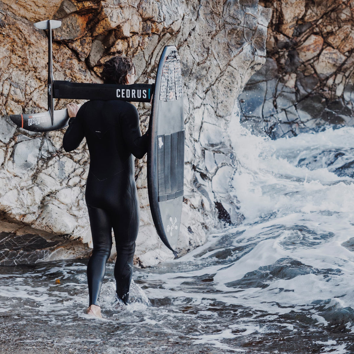 Image with surfer walking out to the ocean against a large cliff. Cedrus masts are custom made, high performance masts for any foil system. 